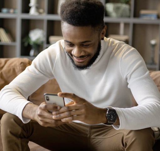 An individual sitting on a brown couch, using a mobile phone and texting, with shelves containing various items in the background.