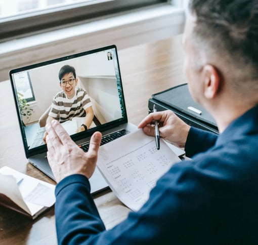 A person engaged in a video call on a laptop, taking notes during the video call, with papers beside the notebook on a wooden table. And also there is another person on video call showing on the screen of the laptop. 