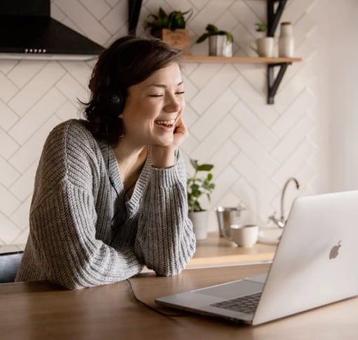 A person in a grey sweater engaged in a video call on a laptop, sitting at a wooden table with a white wall and wooden shelves in the background.